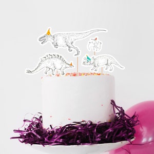 May include: A white cake with purple shredded paper around the base. The cake is decorated with four dinosaur cake toppers, each wearing a party hat. The cake is on a white cake stand.