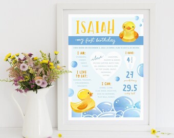 May include: A blue and yellow first birthday poster for a baby boy named Isaiah. The poster features a cartoon rubber ducky, bubbles, and details about the baby's birth, including his weight, height, and favorite things. The text reads "Isaiah, my first birthday, I was born on December 4, 2013, 8:54 PM, 7 lbs 15.4 oz & 20 inches. I am: stubborn, cheeky, sweet, funny. I love: laughing, mummy & daddy, my dog Jupiter, bath time, all animals, Nan & Pop, my toy, music, swimming, sharing. I like to eat: chicken, yoghurt, strawberries, spaghetti. I can: stand, climb, clap, wave, high five, kick, swim, play ball, dance. I have: 4 teeth, 27 pounds, 29.5 inches tall."