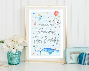May include: A blue and white welcome sign with a watercolor illustration of a whale, shark, and other sea creatures. The sign says "Welcome to Alexander's First Birthday".