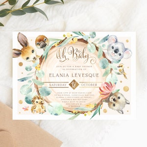 May include: A baby shower invitation featuring a watercolour illustration of Australian animals, including a koala, a kangaroo, a wombat, and an echidna. The invitation is decorated with eucalyptus leaves, flowers, and gold glitter. The text on the invitation reads "Oh Baby! Join us for a baby shower in celebration of Elania Levesque. Saturday, 18 October. 12pm at Bush Bariel Cafe, 123 Main Road, Sunnyville QLD 3033. Elania is registered at Target, Amazon & Buy Buy Baby. Kindly RSVP to Jessie 0412 345-678 by 10 October."