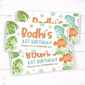 May include: Three white chocolate bar wrappers with a dinosaur theme. The wrappers feature green and orange dinosaurs, green leaves, and colorful polka dots. The text on the wrappers reads "Bodhi's 1st Birthday Thanks for STOMPING by!"