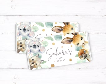 Può includere: Due involucri personalizzati per barrette di cioccolato con un design ad acquerello che presenta un koala, un canguro e un ornitorinco. Gli involucri sono decorati con foglie di eucalipto, punti di glitter dorati e il testo "Sahara's First Birthday".