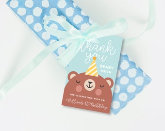 May include: A blue and white polka dot gift bag with a light blue ribbon tied around it. A thank you tag with a brown bear wearing a yellow and white party hat is attached to the ribbon. The tag says "Thank you Beary Much" and "For celebrating with us! Williams 1st Birthday".