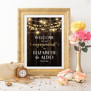 May include: A rustic wooden sign with string lights, announcing "Welcome to the engagement of Elizabeth & Aldo 18.02.22". The sign is framed in gold and displayed on a table with gold accents.