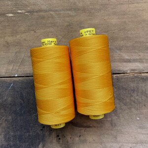 May include: Two spools of bright yellow polyester thread, labeled "Mann Mara", sit on a weathered wooden surface. The thread is tightly wound, and the spools have yellow and white labels. The background is a rustic, dark wood.