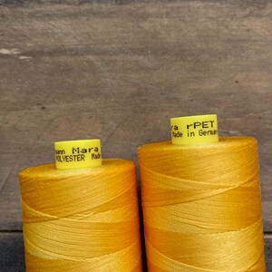 May include: Two spools of bright yellow thread, labeled "Mara Polyester Made in Germany" and "Mara rPET Made in Germany." The thread is tightly wound, and the spools are set against a wooden background. The thread appears to be for sewing or crafting.