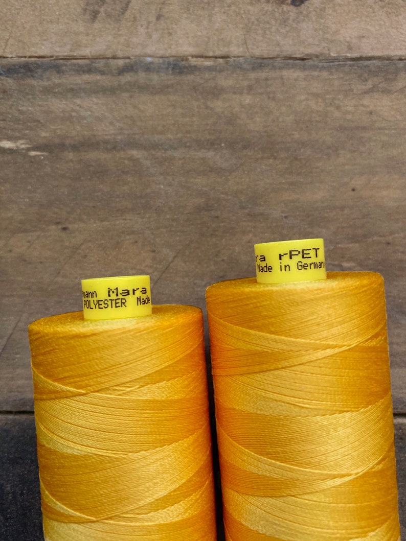 May include: Two spools of bright yellow thread, labeled "Mara Polyester Made in Germany" and "Mara rPET Made in Germany." The thread is tightly wound, and the spools are set against a wooden background. The thread appears to be for sewing or crafting.