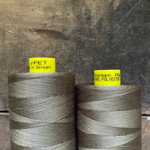 Puede incluir: Dos bobinas de hilo verde oliva. La bobina de la izquierda tiene una etiqueta amarilla que dice "rPET in Germany". La bobina de la derecha tiene una etiqueta amarilla que dice "G&uuml;termann Made in Germany 100% POLYESTER".