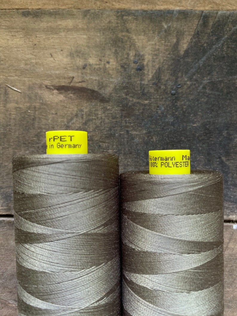 Puede incluir: Dos bobinas de hilo verde oliva. La bobina de la izquierda tiene una etiqueta amarilla que dice "rPET in Germany". La bobina de la derecha tiene una etiqueta amarilla que dice "G&uuml;termann Made in Germany 100% POLYESTER".