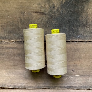 May include: Two spools of beige thread are displayed against a weathered wooden background. The thread is tightly wound on yellow spools. One spool has the number 169 printed on it. The other spool has the text "r-PET Made in German" printed on it.