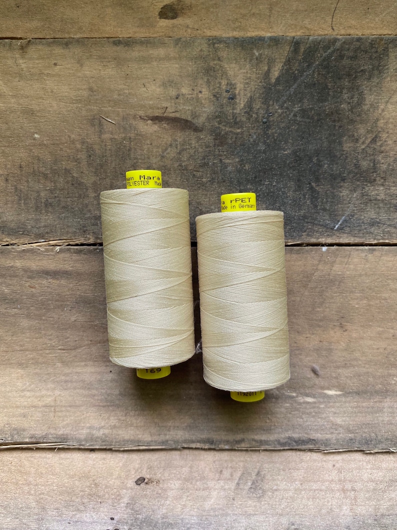 May include: Two spools of beige thread are displayed against a weathered wooden background. The thread is tightly wound on yellow spools. One spool has the number 169 printed on it. The other spool has the text "r-PET Made in German" printed on it.