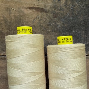 May include: Two spools of cream-colored thread. The spools have yellow labels with black text that reads "Mara POLYESTER Made" and "r-PET Made in Germany". The thread is tightly wound around the spools.