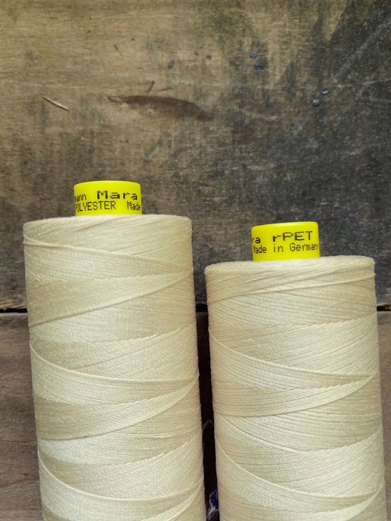 May include: Two spools of cream-colored thread. The spools have yellow labels with black text that reads "Mara POLYESTER Made" and "r-PET Made in Germany". The thread is tightly wound around the spools.