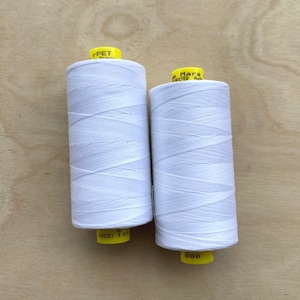 May include: Two spools of white thread are displayed against a light wood grain background. The thread is tightly wound around the spools, and the labels are yellow. One spool has the text "rPET" on the label.