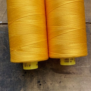 May include: Two spools of bright yellow thread. Each spool has a yellow label with the text "Col. 417" and "1093 yds 1000 m". The thread is tightly wound and has a slight sheen. The background is a dark, textured surface.