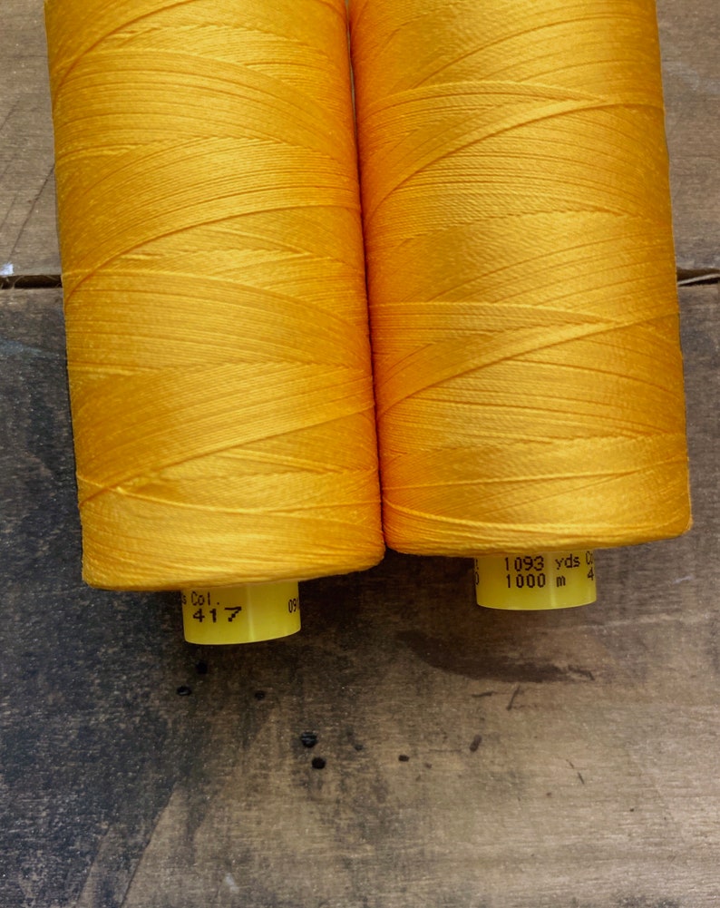 May include: Two spools of bright yellow thread. Each spool has a yellow label with the text "Col. 417" and "1093 yds 1000 m". The thread is tightly wound and has a slight sheen. The background is a dark, textured surface.