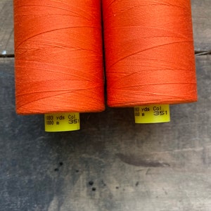 May include: Two spools of orange thread are displayed against a weathered wooden surface. Each spool is tightly wound with vibrant thread, and the labels indicate 1093 yards (999.7 meters) of thread. The color code is 351.