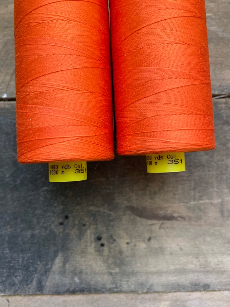 May include: Two spools of orange thread are displayed against a weathered wooden surface. Each spool is tightly wound with vibrant thread, and the labels indicate 1093 yards (999.7 meters) of thread. The color code is 351.