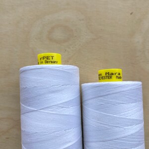 May include: Two spools of white thread with yellow labels. The left spool has the text "rPET in Germany" and the right spool has the text "Mara POLYESTER Made". The spools are on a light brown wooden surface.