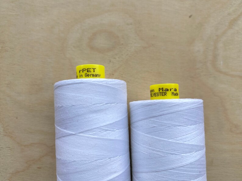 May include: Two spools of white thread with yellow labels. The left spool has the text "rPET in Germany" and the right spool has the text "Mara POLYESTER Made". The spools are on a light brown wooden surface.