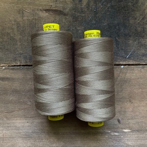 Puede incluir: Dos bobinas de hilo marr&oacute;n con etiquetas amarillas. Las etiquetas dicen "rPET in Germany" y "G&uuml;termann Made in 100% POLYESTER". El hilo est&aacute; enrollado apretadamente alrededor de las bobinas. Las bobinas est&aacute;n sobre un fondo de madera.