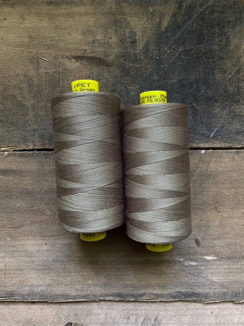 Puede incluir: Dos bobinas de hilo marr&oacute;n con etiquetas amarillas. Las etiquetas dicen "rPET in Germany" y "G&uuml;termann Made in 100% POLYESTER". El hilo est&aacute; enrollado apretadamente alrededor de las bobinas. Las bobinas est&aacute;n sobre un fondo de madera.