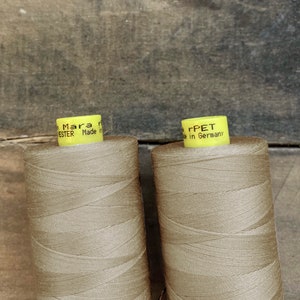 K&ouml;nnte beinhalten: Zwei Spulen beigefarbenes Garn mit gelben Etiketten. Auf den Etiketten steht "Mara" und "Polyester Made in Germany". Das Garn ist eng auf die Spulen gewickelt, vor einem verwitterten Holzgrund.