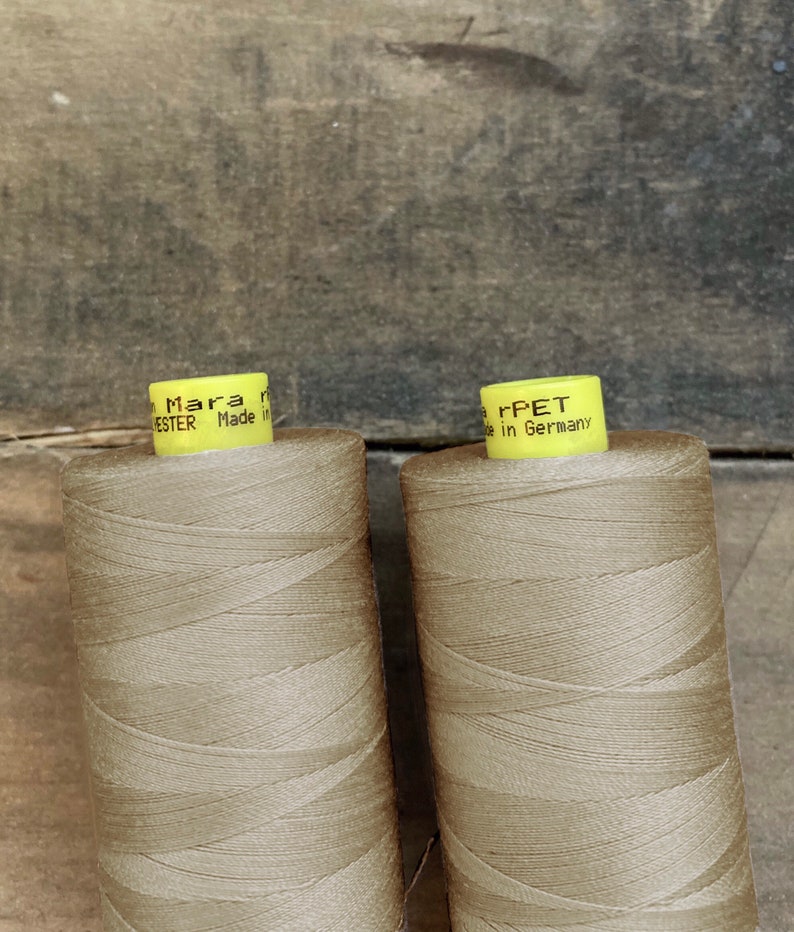 K&ouml;nnte beinhalten: Zwei Spulen beigefarbenes Garn mit gelben Etiketten. Auf den Etiketten steht "Mara" und "Polyester Made in Germany". Das Garn ist eng auf die Spulen gewickelt, vor einem verwitterten Holzgrund.