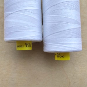 May include: Two spools of white thread are displayed against a light wood background. The thread is tightly wound around the spools. Each spool has a yellow label with black text. One label reads "1642021 1 00" and the other reads "Col. 800".