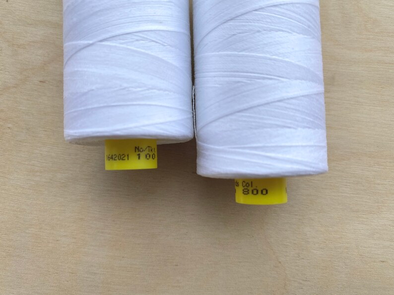 May include: Two spools of white thread are displayed against a light wood background. The thread is tightly wound around the spools. Each spool has a yellow label with black text. One label reads "1642021 1 00" and the other reads "Col. 800".