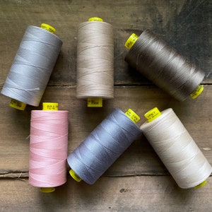 Puede incluir: Una colecci&oacute;n de seis carretes de hilo en varios colores neutros, incluyendo gris, beige y marr&oacute;n. Los carretes est&aacute;n dispuestos sobre una superficie de madera r&uacute;stica, con etiquetas amarillas visibles. El hilo parece estar bien enrollado.