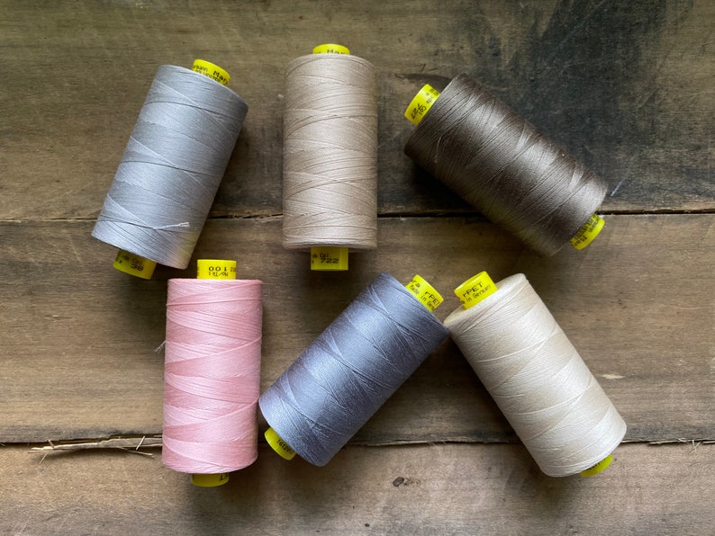 Puede incluir: Una colecci&oacute;n de seis carretes de hilo en varios colores neutros, incluyendo gris, beige y marr&oacute;n. Los carretes est&aacute;n dispuestos sobre una superficie de madera r&uacute;stica, con etiquetas amarillas visibles. El hilo parece estar bien enrollado.