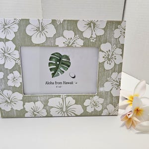 May include: A green and white picture frame with a floral design. The frame has a white mat and a clear glass front. The frame is 4x6 inches and says "Aloha from Hawaii".