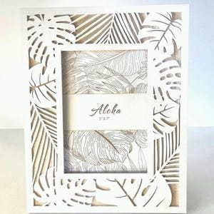 May include: White wooden picture frame with a cutout design of tropical leaves. The frame is for a 5x7 inch photo and has the word "Aloha" printed on it.