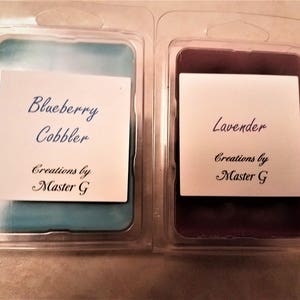 May include: Two wax melts in clear plastic containers. The left melt is blue and says "Blueberry Cobbler" and "Creations by Master G". The right melt is purple and says "Lavender" and "Creations by Master G".