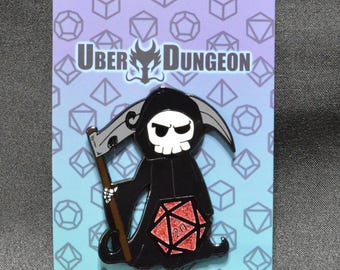 May include: A black enamel pin featuring a Grim Reaper character with a scythe and a red and black D20 die. The pin is on a card with the text "Uber Dungeon" and a geometric pattern in blue and purple.