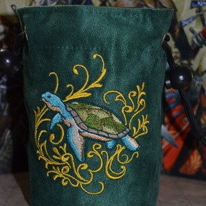 May include: Dark green drawstring bag with an embroidered sea turtle design. The turtle is blue, green, and brown, surrounded by gold filigree. The bag is made of a soft, velvety material.