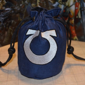 May include: A navy blue drawstring dice bag with a silver embroidered symbol. The bag is round and has a black drawstring closure. The symbol is a stylized letter or emblem.