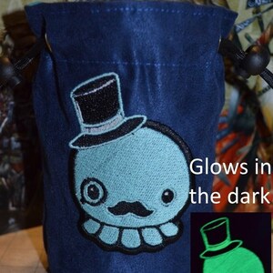 May include: A dark blue drawstring pouch with a light blue embroidered cartoon character wearing a top hat and moustache. The text "Glows in the dark" is visible. A smaller image shows the character glowing green.