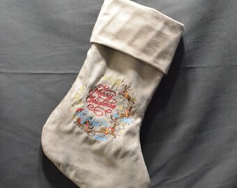 May include: A beige Christmas stocking with a folded cuff, hanging against a gray background. The stocking features an embroidered design with the words "Merry Christmas" and a festive scene with reindeer and a sleigh.
