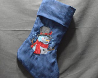 May include: A blue Christmas stocking with a snowman design. The snowman has a black top hat, a red scarf, and twig arms. The stocking is made of a soft, velvety material and is hanging from a hook.