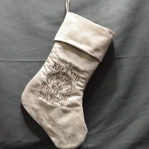 May include: A light beige Christmas stocking with a dark brown embroidered design. The design includes a dragon, sword, and the text "What doesn't kill you gives you XP". The stocking is hanging from a silver hook.