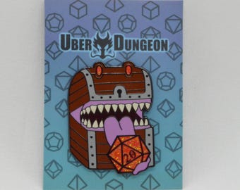 May include: A pin featuring a cartoon treasure chest with a large mouth and tongue, holding a glittery orange D20 die. The pin has the text "UBER DUNGEON" at the top, set against a blue background with geometric shapes.
