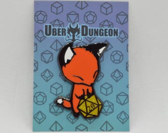 May include: A pin featuring a cartoon fox with orange fur, black ears, and a white face, holding a yellow twenty-sided die. The pin is on a blue card with the text "UBER DUNGEON" and a geometric pattern.