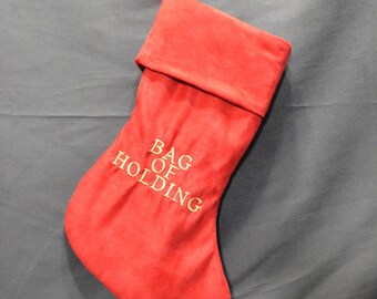 May include: Red Christmas stocking with the words "BAG OF HOLDING" embroidered in gold thread. The stocking is hanging from a silver hook. The stocking is made of a soft, velvety material.