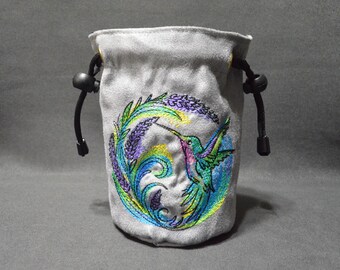 May include: A gray drawstring pouch with a colorful embroidered hummingbird design. The hummingbird is depicted with green, blue, and pink hues, surrounded by a circular pattern of leaves and lavender. Black drawstrings and toggles complete the design.