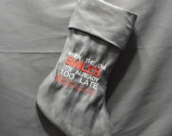 May include: Gray Christmas stocking with the text "WHEN THE DM SMILES IT'S ALREADY TOO LATE" embroidered in red. The stocking is hanging from a silver hook. The background is a gray fabric.