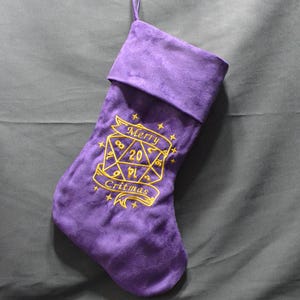 May include: A purple Christmas stocking with a gold embroidered design featuring a D20 die and the words "Merry Critmas." The stocking is made of a soft material and has a loop for hanging.