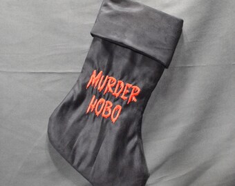 May include: A dark gray Christmas stocking with the words "MURDER HOBO" embroidered in orange. The stocking is hanging from a metal hook against a gray background. The top of the stocking is folded over.