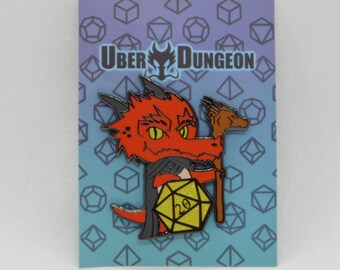 May include: A red dragon enamel pin with a yellow twenty-sided die and a brown staff. The pin is on a blue card with geometric shapes and the text "UBER DUNGEON". The dragon has yellow eyes and is wearing a black cloak.